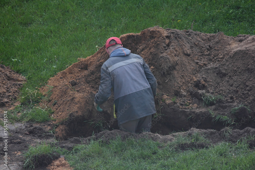 One European digger worker in gray overalls stands with a shovel up to his waist in a ditch on a summer day against a background of green grass, local landscaping works