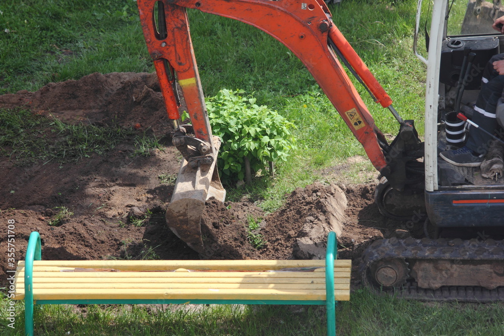 A compact crawler excavator on rubber tracks carefully digs a ditch with a bucket in the Park on a summer day against the background of green grass, a Bush and a bench, local landscaping works