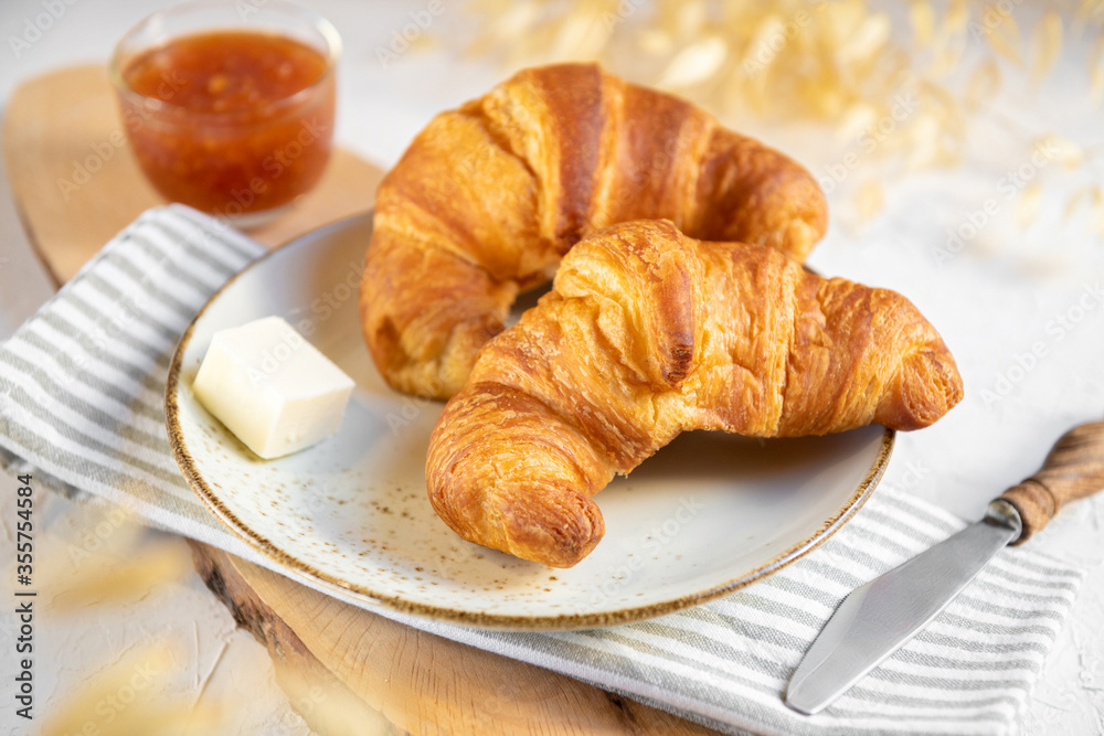 A pair of croissants on ceramic plate with butter and orange jam on a wooden cutten board. Continental breakfast background, close up