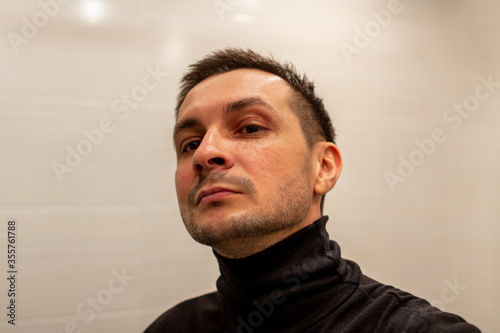 Portrait of a young short haired man wearing black turtleneck on background of light and looking looking forward, head bowed.