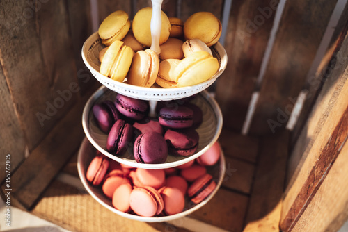 Three tiered plate stand with colorful sweet macaroons. Wooden plant box background. Dessert for served with afternoon tea or coffee. Wedding or party