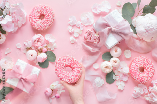 Top view composition of hand holds donut and two more icing donuts, two gift boxes with silk pink ribbons and bows and pink flowers on a pastel pink background. Festive concept.