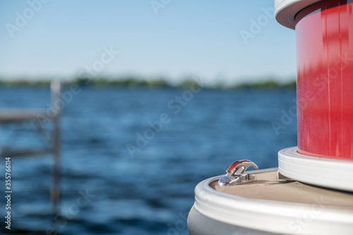 Detail of a metal cleat on the front of a red pontoon boat with a dock, lake and horizon in the background, with selective focus and narrow depth of field.