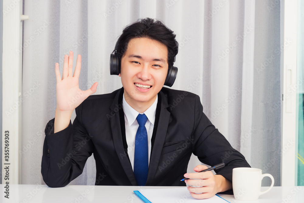 Work from home, Asian young businessman wear suit video conference call or facetime he smiling sitting on desk wearing headphones and raise your hand to say hello greet colleague at home office Work from home, Asian young businessman wear suit video conference call or facetime he smiling sitting on desk wearing headphones and raise your hand to say hello greet colleague at home office
