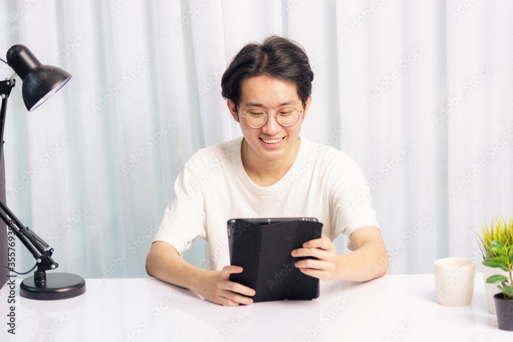 Happy Asian young business handsome man work from home office wear glasses, t-shirt comfortable he smiling and using a black modern smart digital tablet computer to read email or learning on desk Happy Asian young business handsome man work from home office wear glasses, t-shirt comfortable he smiling and using a black modern smart digital tablet computer to read email or learning on desk