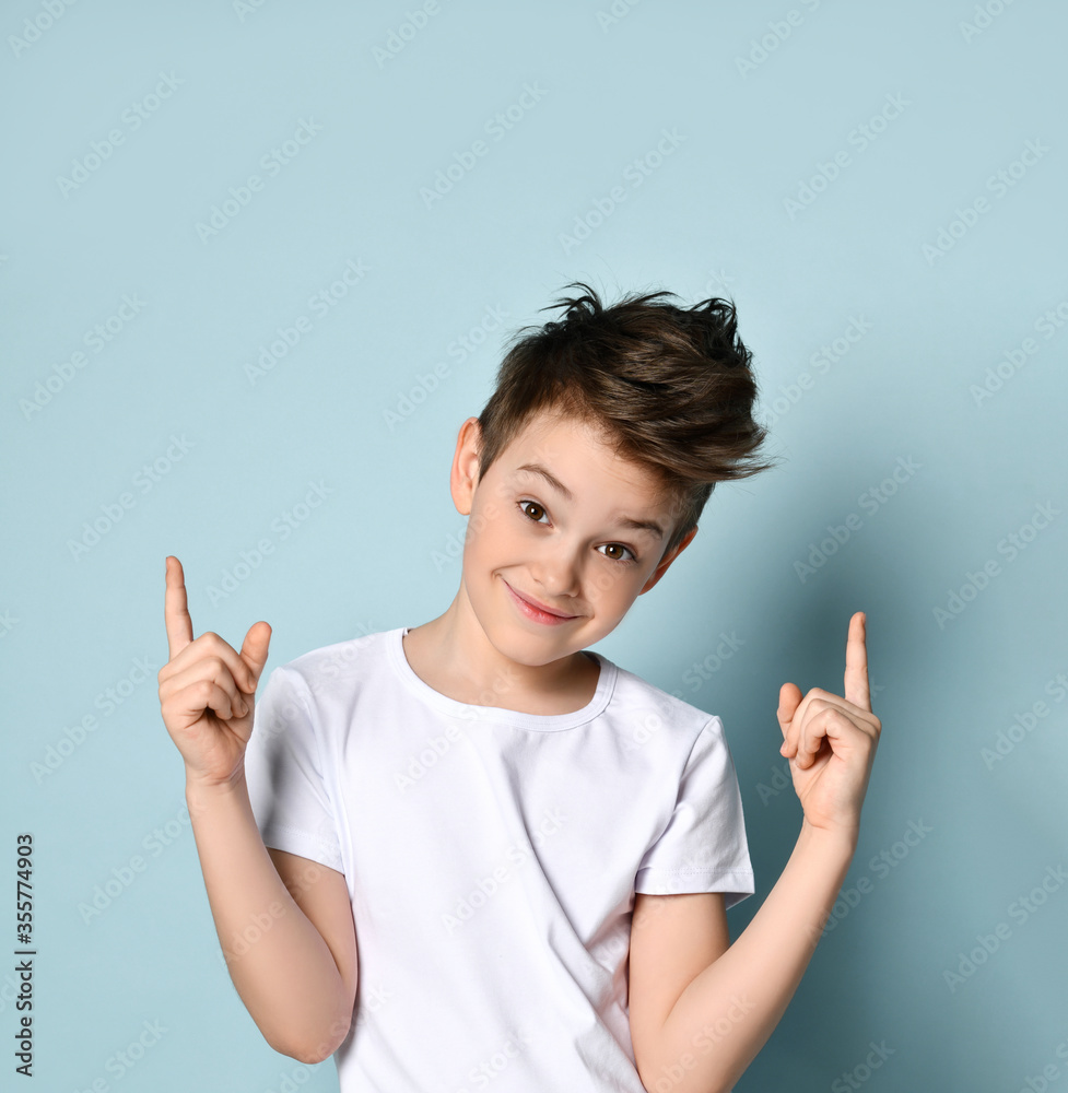 Naughty kid looking at camera with broad sly smile pointing his index fingers up getting up to mischief. Half-length portrait isolated on light blue Naughty kid looking at camera with broad sly smile pointing his index fingers up getting up to mischief. Half-length portrait isolated on light blue