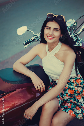 Portrait of a young brunette in a colorful dress on a summer scooter in the city 