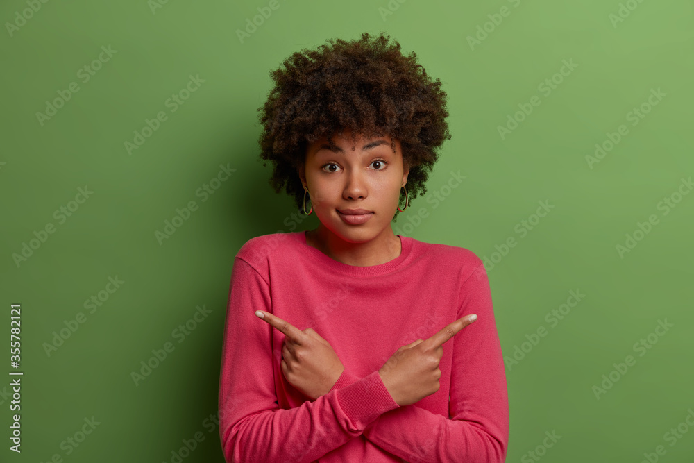 Beautiful perplexed dark skinned woman has hard complicated choice, points index fingers sideways at two objects, crosses hands, should decide what to choose, dressed in bright clothes, poses indoor Beautiful perplexed dark skinned woman has hard complicated choice, points index fingers sideways at two objects, crosses hands, should decide what to choose, dressed in bright clothes, poses indoor