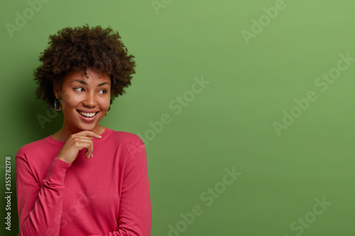 Dreamy cheerful curly African American woman holds hand on chin, sees something pleasant and appealing, dressed in pink jumper, stands against green background, free space for your advertisement