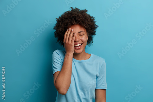 Horizontal shot of happy dark skinned woman makes face palm, giggles positively, keeps eyes closed, has good mood, wears blue t shirt, poses indoor, hears amazing info, reacts to lucky fine situation