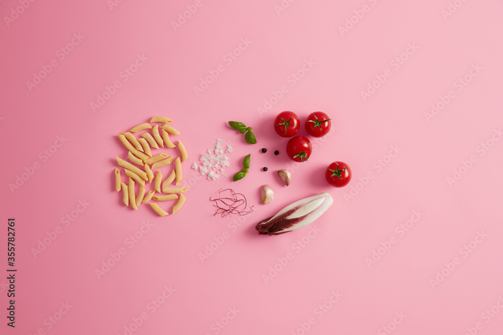 Penne dry rice pasta basil, chicory salad, tomatoes, garlic red chili pepper threads for preparing delicious gourmet Italian cuisine. Uncooked macaroni and ingredients on rosy background. Healthy food