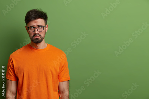 Unhappy gloomy young European man looks upset and disappointed, wears casual orange t shirt and spectacles, feels uneasy and moody, stands against green background, copy space for your promotion.