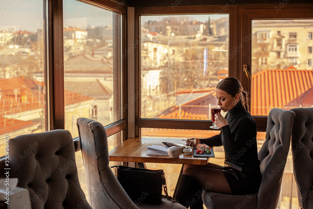 Female posing in cafe on the roof of the city and using a laptop. Fashion woman dressed style black golf and shorts. Freelancer relexing on the rooftop and holding computer, eating sushi. Female posing in cafe on the roof of the city and using a laptop. Fashion woman dressed style black golf and shorts. Freelancer relexing on the rooftop and holding computer, eating sushi.