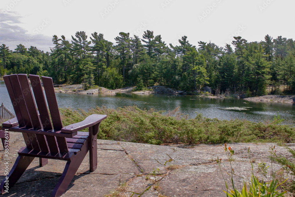 Fototapeta premium Two black Muskoka chairs sitting on a wood dock facing a lake. Across the calm water is a white cottage nestled among green trees. Canada flag is visible