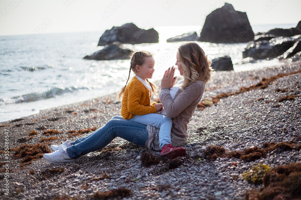 Happy family posing outdoor in the beach of the sea at spring time. woman with daughter have fun on vacation near ocean. Female parent holds the child and tickles. 
