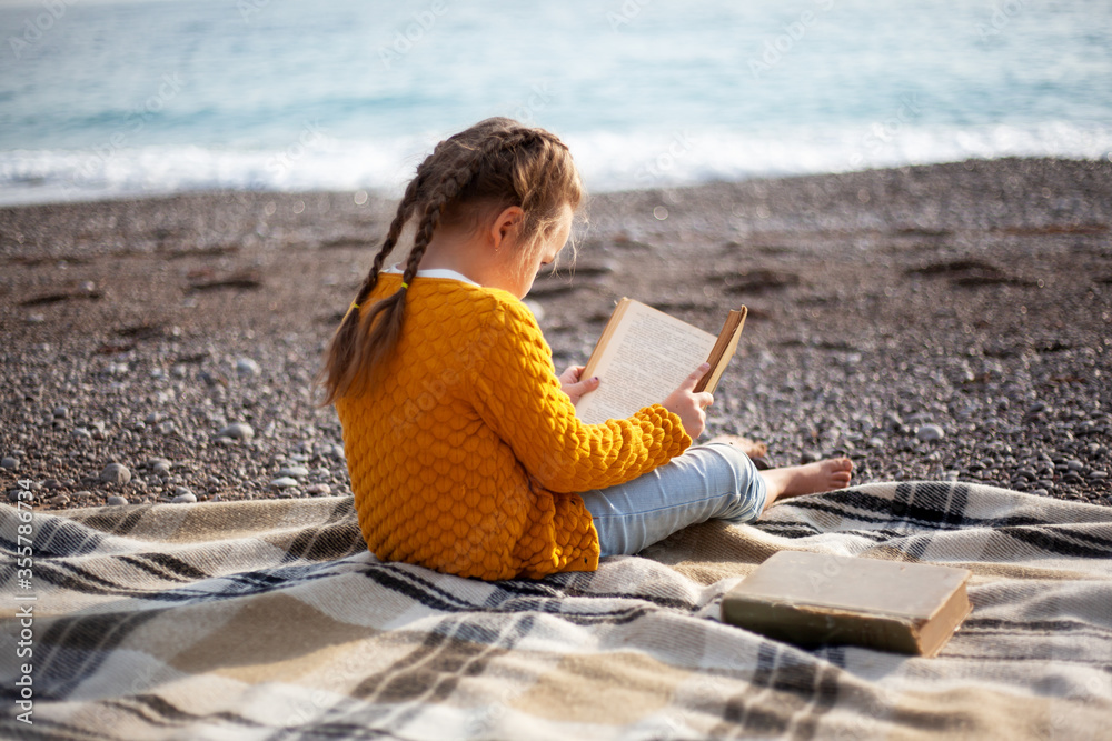 Female kid read the book and relax in the blanket in the beach against stone. Barefoot child relax on the sand at the shore of sea in the nature . Happy leisure have fun vacation