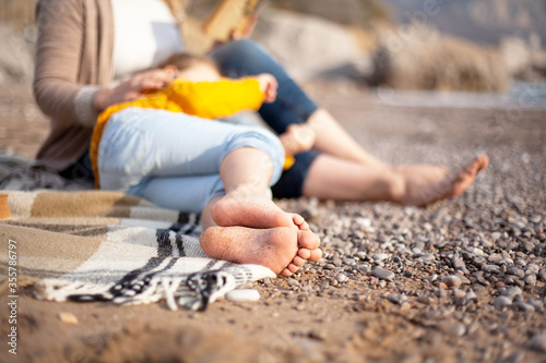 Close-up of barefoot heels of kid girl. Woman read the book and relax in the blanket in the beach against stone while kid sleep on the legs. Happy leisure to relax with family on the sea shore