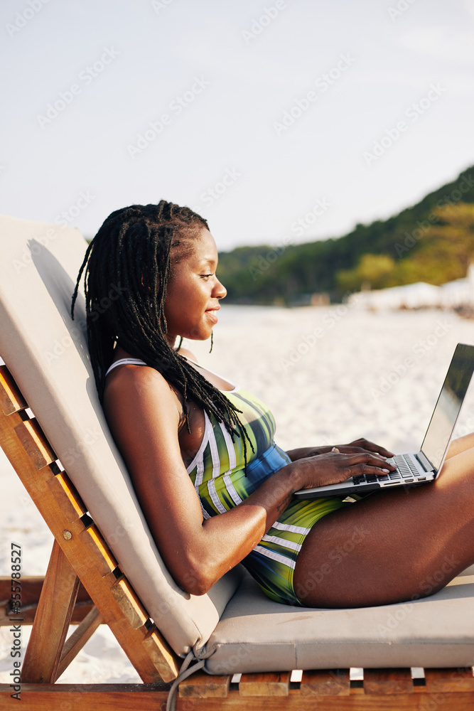 Smiling pretty young Black woman in swimsuit sitting on chaise-lounge and working on laptop