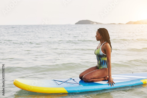 Photography Beautiful fit smiling young Black woman in swimming suit sitting on sup board an