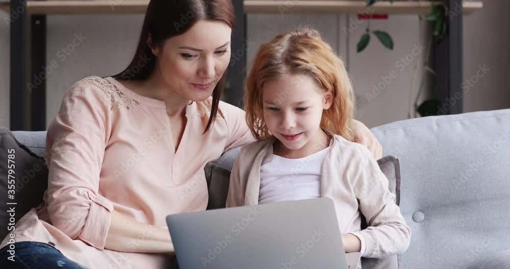 Caring young mum sitting on couch with little redhead daughter, showing educational application on computer, teaching using tech software indoors. Smart small child girl studying online on laptop. Caring young mum sitting on couch with little redhead daughter, showing educational application on computer, teaching using tech software indoors. Smart small child girl studying online on laptop.