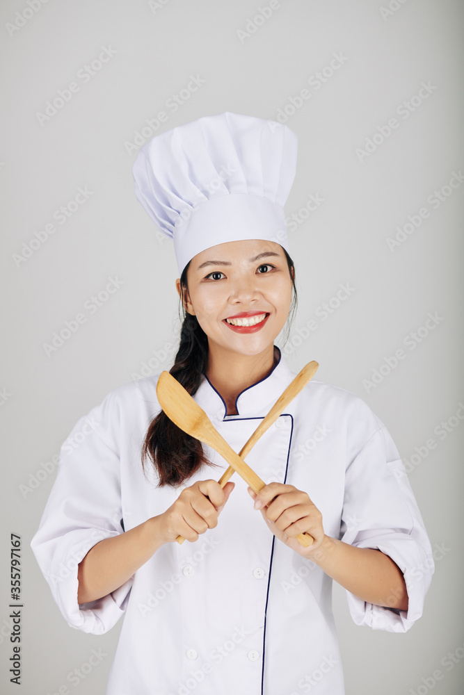 Portrait of pretty smiling young Asian cook crossing wooden spoon and fork Portrait of pretty smiling young Asian cook crossing wooden spoon and fork