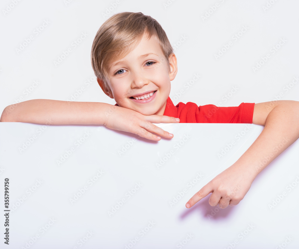 Photo of a happy smiling kid look outs from the blank billboard. Portrait of cheerful child pointing on white banner. Portrait of a 8 years old boy is signing by finger to banner Photo of a happy smiling kid look outs from the blank billboard. Portrait of cheerful child pointing on white banner. Portrait of a 8 years old boy is signing by finger to banner