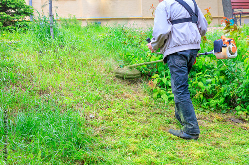 A worker cuts tall grass with an industrial petrol trimmer in hard-to-reach areas of a city lawn. photo