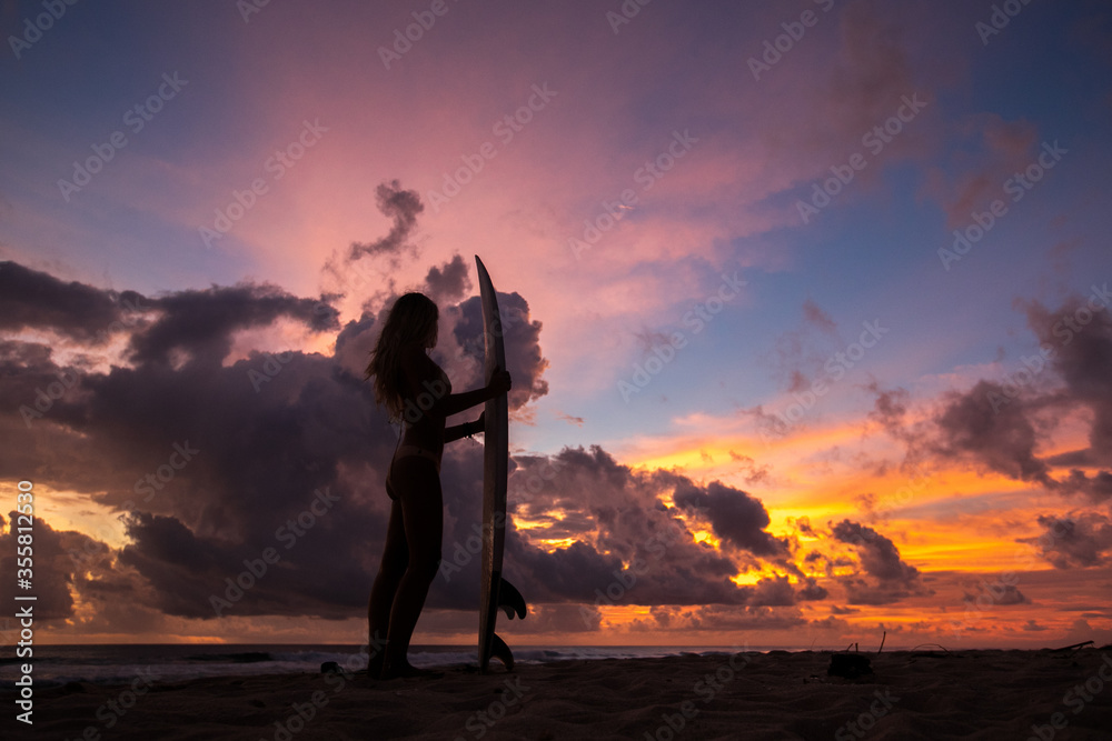 surf girl meets sunset on the beach. silhouette. a young girl with a surfboard stands on the beach and looks at a stunning sunset. multi-colored sky and clouds.