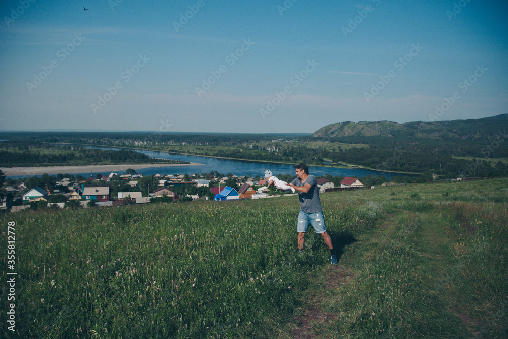 father walks with his daughter in meadow. man throws child. Baby in flight. Village houses, forest and river as background. oncept of summer, warmth, freedom, village life, sunburn, family day