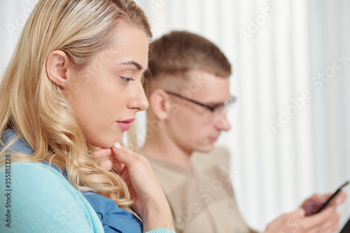 Pensive pretty young woman sitting next to her husband and reading important text message
