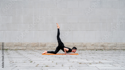 Chaturanga pose with one leg up. Fit latin man do yoga outdoor on orange mat with gray concrete wall at the background. Copy space.