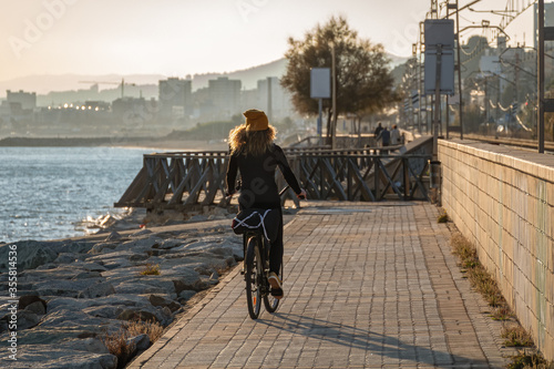 Young curly girl in a yellow hat rides a bicycle at sunset on the promenade road of Maresme, Catalonia, Spain. photo