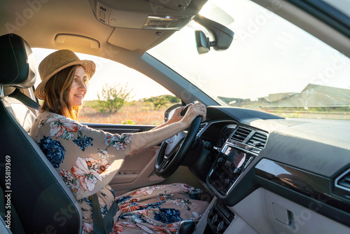 Картината върху платно A smiling woman in a hat and a colored dress is sitting behind the wheel of a ca