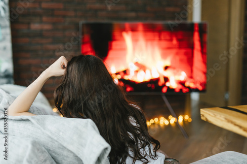 
The girl is resting by the fireplace. Girl is watching TV with the image of fire and fireplace. The girl is resting in the living room.
