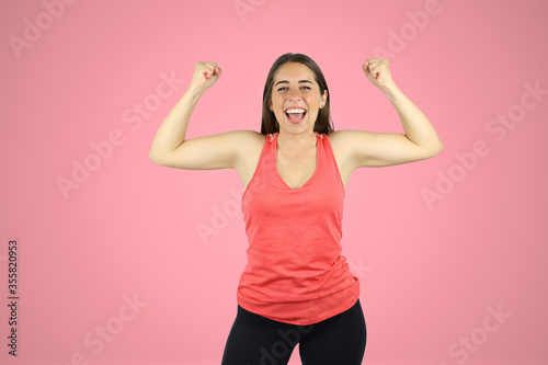 Young beautiful woman standing over pink isolated background wearing sport chlotes and working out. Very happy and smailing with her arms up with victory gesture.