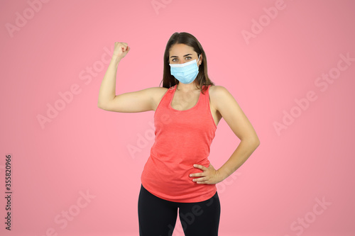 Young beautiful woman standing over pink isolated background wearing sport chlotes and mask. Doing a gesture with his arm of strength and victory