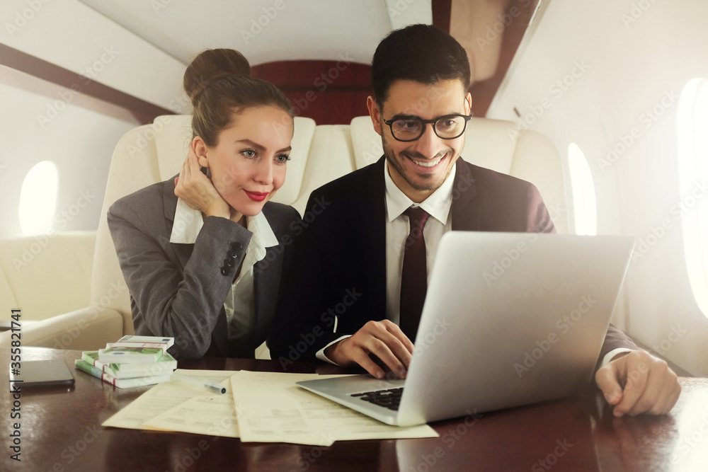 Business man and woman flying first class, sitting at table in airplane with packs of banknotes, looking in open laptop, analysing sales data Business man and woman flying first class, sitting at table in airplane with packs of banknotes, looking in open laptop, analysing sales data