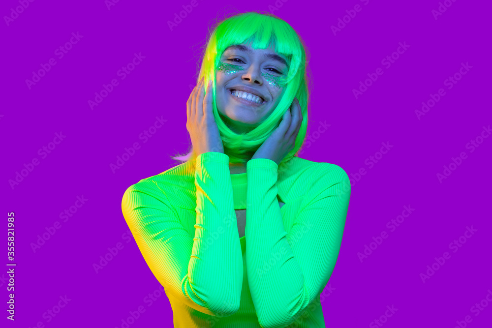 Close-up portrait of young happy teenage girl wearing neon green top and yellow wig, smiling and touching her hair as if feeling shy, isolated on purple background Close-up portrait of young happy teenage girl wearing neon green top and yellow wig, smiling and touching her hair as if feeling shy, isolated on purple background