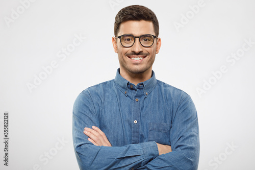 Young business guy wearing blue shirt and eyeglasses, standing with arms crossed, smiling happily and feeling confident, isolated on gray background