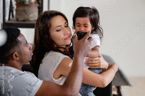 Little mixed race girl crying sitting on the hands of a caring mom and a comforting african-american dad. Concept of child care and the addiction of children to gadgets