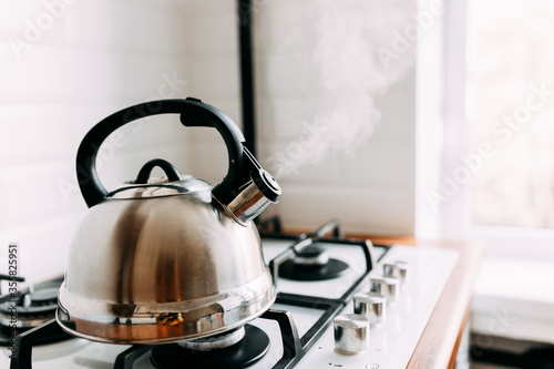 Obraz na płótnie The kettle boils, steam from metal teapot on gas oven. Bright kitchen interior. White modern dining room. Wooden complete kitchen with gas oven. w salonie
