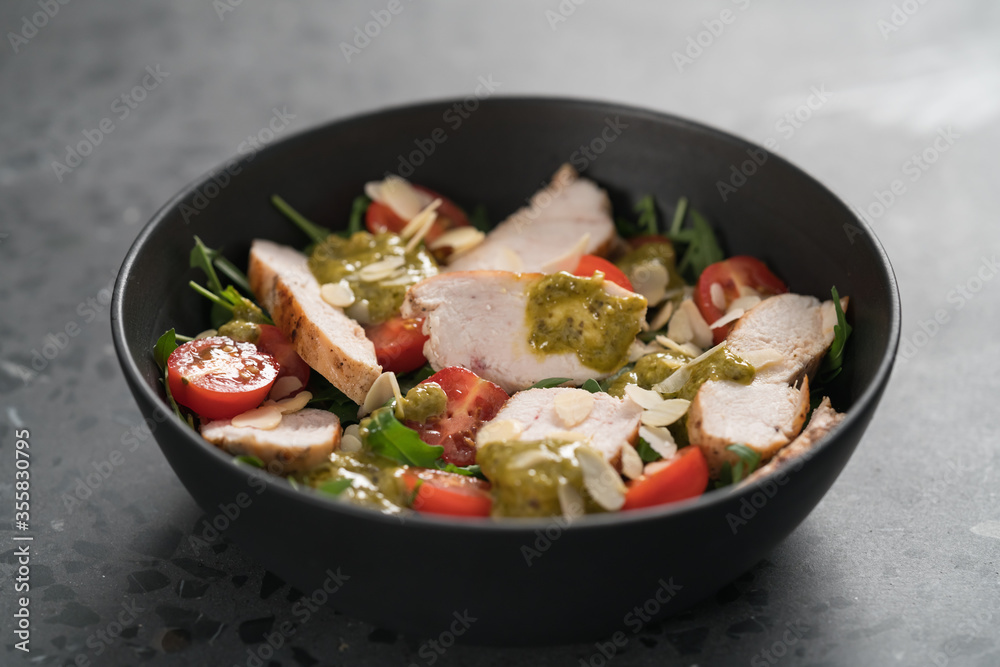 Salad with chicken, tomatoes and arugula in black bowl on concrete background