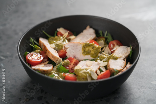 Salad with chicken, tomatoes and arugula in black bowl on concrete background
