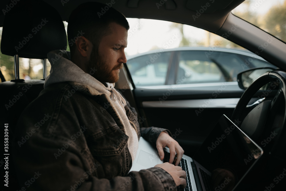 A young man with a beard opens his laptop computer to do business inside a comfort car. A guy stopped his car to immediately remotely solve tasks at work in social distance.