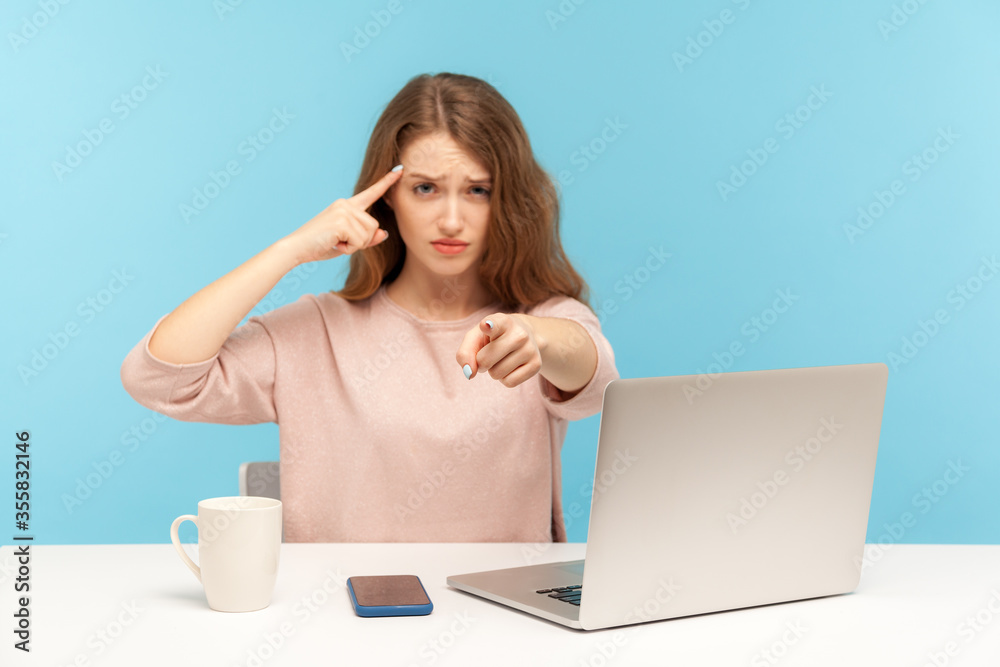 You are idiot! Woman with sarcastic look sitting at workplace and making stupid gesture, pointing to camera, teasing and accusing senseless dumb mind. indoor studio shot isolated on blue background You are idiot! Woman with sarcastic look sitting at workplace and making stupid gesture, pointing to camera, teasing and accusing senseless dumb mind. indoor studio shot isolated on blue background