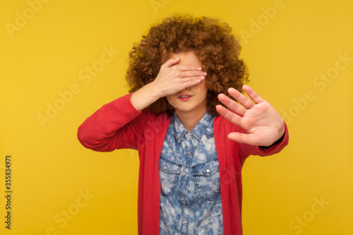 No, don't want to look. Portrait of scared woman with curly hair covering eyes and showing stop gesture, feeling shy afraid to watch, ignoring troubles. studio shot isolated on yellow background