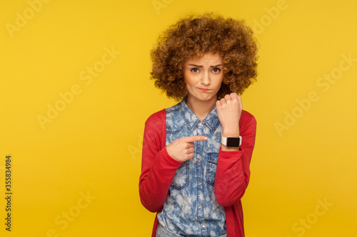 Portrait of upset impatient woman with curly hair showing wrist watch and looking disappointed at camera, reminding of late time, asking to hurry. indoor studio shot isolated on yellow background