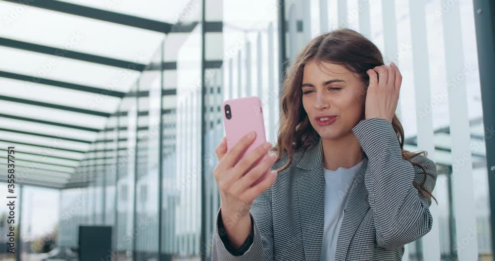 Attractive girl smiling and talking while having video conversation. Millennial businesswoman gesturing and waving while using smartphone app for communication near modern building. Attractive girl smiling and talking while having video conversation. Millennial businesswoman gesturing and waving while using smartphone app for communication near modern building.