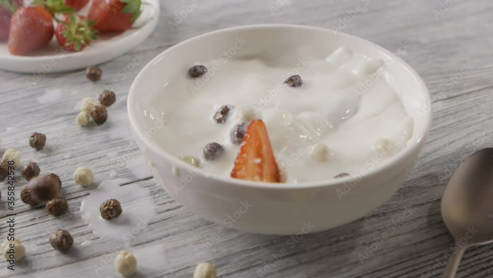 Ripe red strawberry fruit and corn balls fall into a white bowl with milk with a splash. Close-up, slow motion. The milk splash falls out of the bowl on the gray table. Full HD video, 240fps,1080p. Ripe red strawberry fruit and corn balls fall into a white bowl with milk with a splash. Close-up, slow motion. The milk splash falls out of the bowl on the gray table. Full HD video, 240fps,1080p.