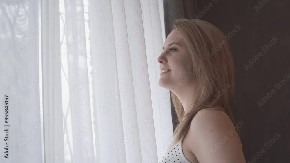 Beautiful young european woman is awake and walking to a window with an open curtain in a bedroom. Sexy girl smiling in underwear is looking out the window, lifestyle concept Beautiful young european woman is awake and walking to a window with an open curtain in a bedroom. Sexy girl smiling in underwear is looking out the window, lifestyle concept